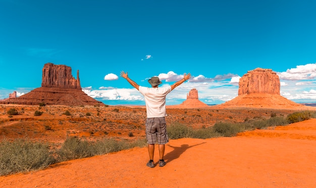 Homme en t-shirt blanc et chapeau vert avec les bras levés dans le parc national de Monument Valley à The Mittens