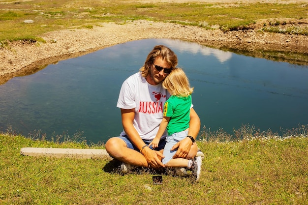 un homme en T-shirt blanc embrasse son petit fils sur le fond d'un petit lac dans les montagnes