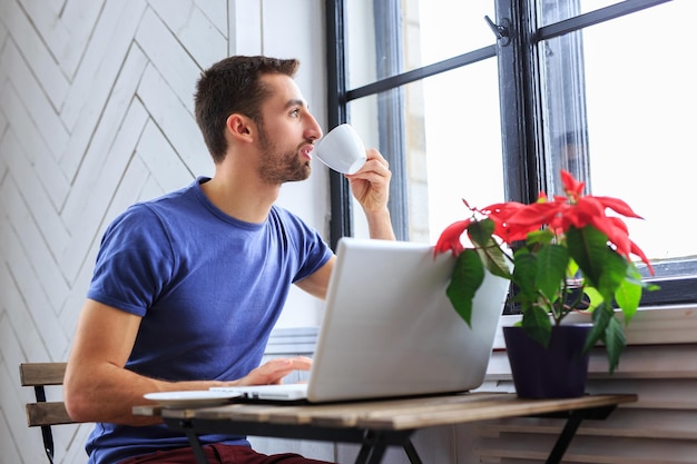 Un homme en t-shirt bleu boit du café et travaille avec un ordinateur portable.