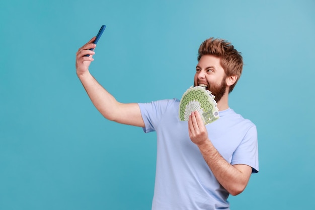 Homme en T-shirt bleu faisant du selfie avec un grand fan de billets en euros montrant de l'argent aux abonnés