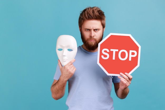 Homme en T-shirt bleu tenant un panneau d'arrêt rouge et un masque blanc regardant la caméra avec une expression stricte