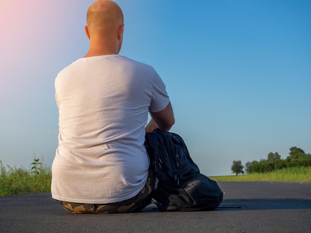 Un homme en T-shirt est assis dos à la caméra sur une route goudronnée et regarde au loin, un sac à dos noir est allongé à côté de lui. Concept de voyage