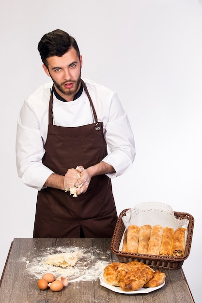 Un homme en tablier prépare la pâte pour la cuisson, modèle Cook sur un mur blanc pétrit la pâte sur la table décorée de produits de boulangerie