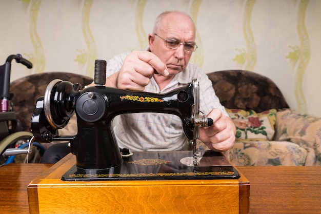 Photo homme de tailleur senior sérieux avec tête chauve, mettant du fil sur sa machine à coudre manuelle à l'intérieur de sa maison.