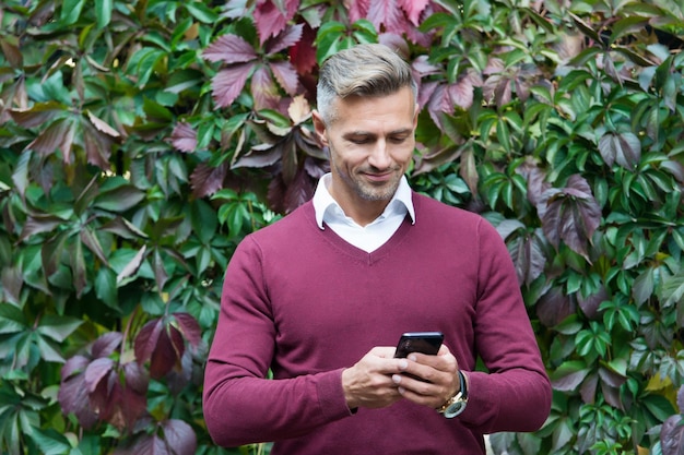 Homme tapant un message homme charismatique et réussi en plein air beau mâle utilisant un téléphone portable moderne mec mûr sur les feuilles d'automne fond automne affaires mode décontractée