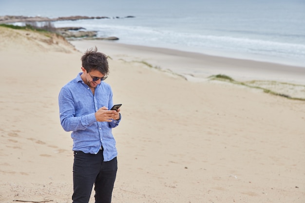 Homme tapant sur son téléphone intelligent en se tenant debout sur la plage tandis qu'en arrière-plan, vous pouvez voir la mer, le sable et les belles vagues.