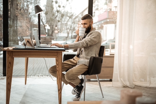 Homme avec tasse. Bel homme barbu attentif en cardigan tricoté en sirotant une boisson chaude tout en restant dans l'armoire