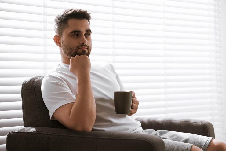 Photo homme avec une tasse de boisson assis sur un fauteuil près des stores de fenêtre à l'intérieur