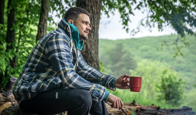 Un homme avec une tasse de boisson chaude se réchauffe au coin du feu dans la forêt.