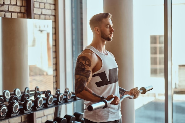 Un homme tatoué musclé fait ses exercices avec une barre dans la salle de sport près de la fenêtre. Miroir et haltères à l'arrière-plan.