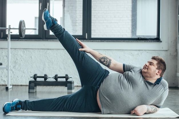 Homme tatoué en surpoids allongé sur un tapis de fitness et étirant les jambes au gymnase