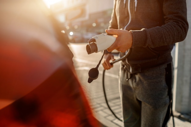 Homme tenant le câble de charge d'alimentation pour voiture électrique dans un parking extérieur. Et il va connecter la voiture à la borne de recharge dans le parking près du centre commercial