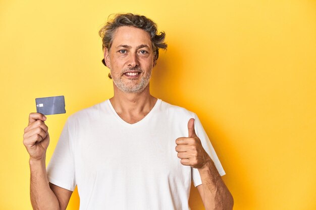 Photo un homme tenant une carte de crédit, un décor de studio jaune, souriant et levant le pouce.