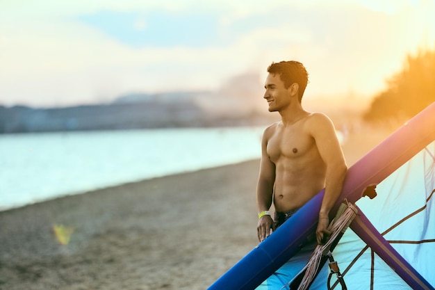 Homme tenant un cerf-volant dans les mains debout sur la plage au coucher du soleil après une bonne séance de kitesurf