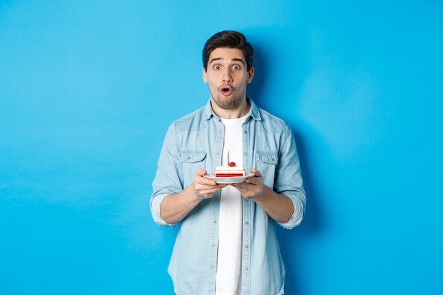 Homme tenant un gâteau d'anniversaire et ayant l'air surpris, faisant un vœu sur une bougie allumée, debout sur fond bleu.