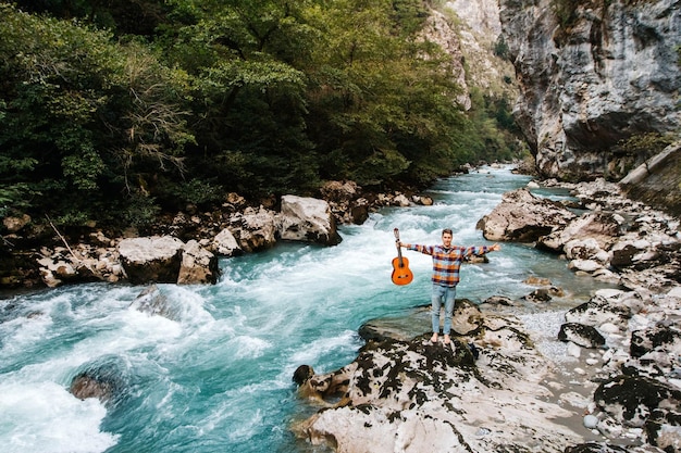 Homme tenant une guitare debout sur la rive d'une rivière de montagne