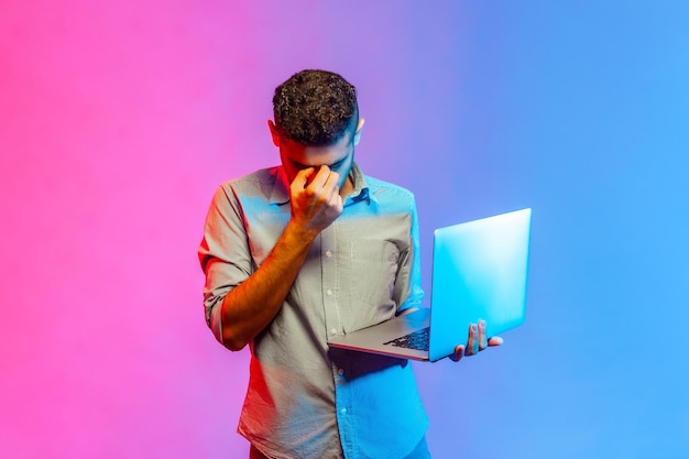 Homme tenant un ordinateur portable dans les mains et se frottant les yeux étant épuisé de travailler de longues heures