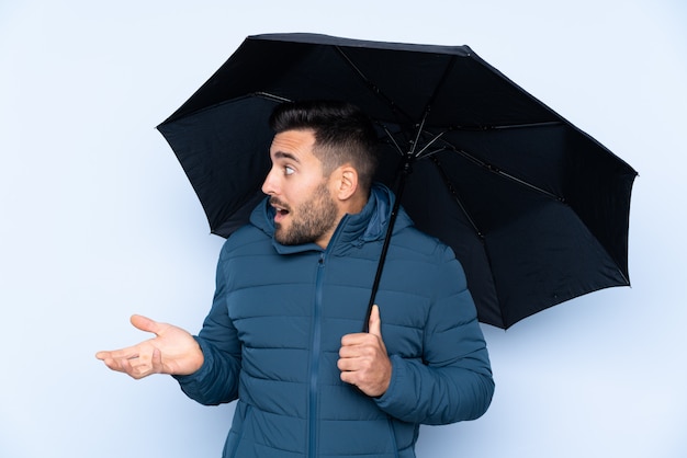 Homme tenant un parapluie sur un mur isolé avec une expression faciale surprise