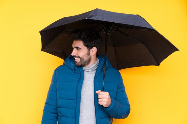 Homme tenant un parapluie sur un mur jaune isolé avec une expression heureuse