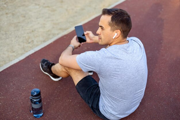 Homme tenant un smartphone dans ses mains assis sur un terrain de sport public