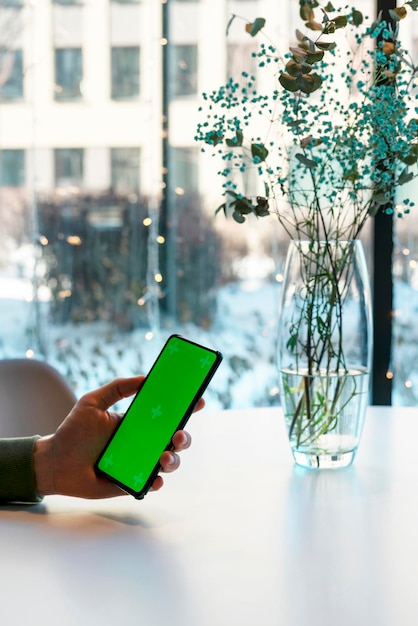 Homme tenant un smartphone avec écran Chroma Key dans un café à la maquette d'entreprise d'écran vert de table blanche