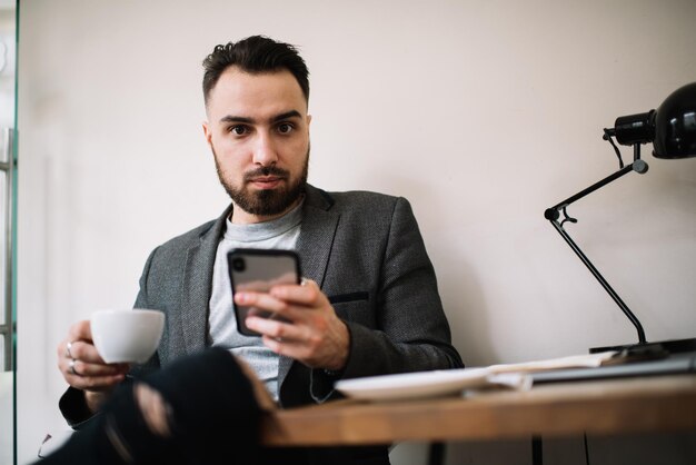 Homme tenant une tasse et un smartphone et regardant la caméra à table au bureau