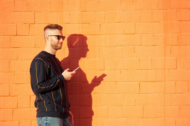 Un homme tenant un téléphone alors qu'il se tient contre un mur de briques orange