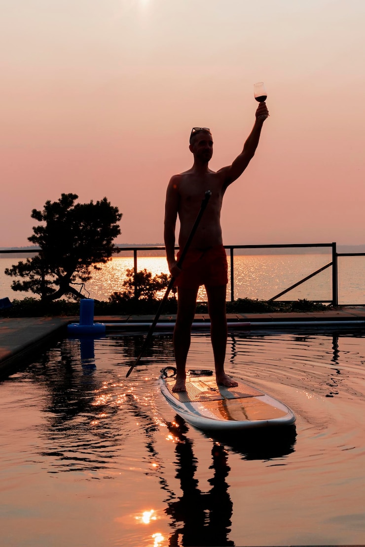Photo un homme tenant un verre de vin alors qu'il se tient sur un paddleboard dans la piscine au coucher du soleil