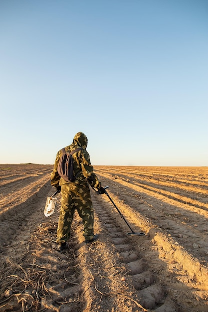 Un homme en tenue de camouflage avec un sac à dos, un détecteur de métaux et une pelle traverse le champ