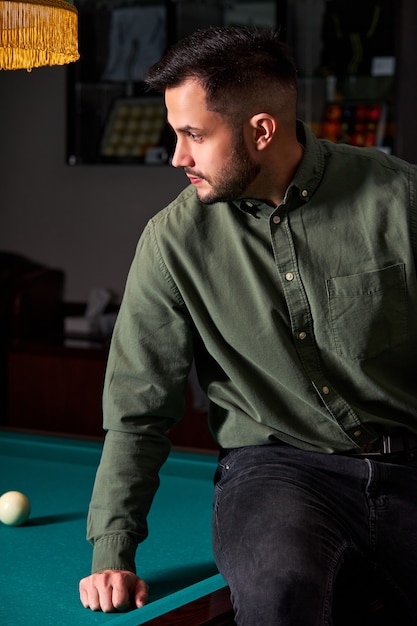 Homme en tenue décontractée est assis sur une table de billard, regardant le jeu, concept de jeu de sport de snooker. portrait