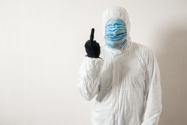 Un homme en tenue de protection accroché avec des masques médicaux posant sur un fond de mur montrant divers gestes avec ses doigts le scientifique montre le majeur