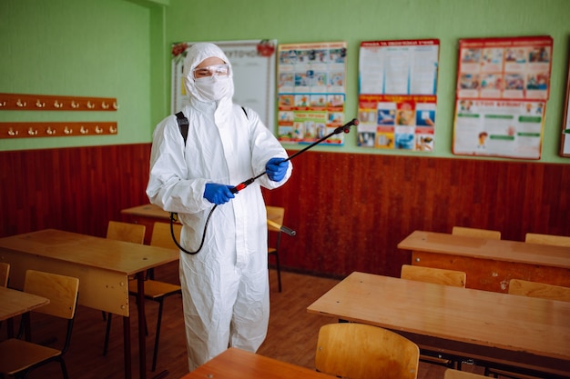 Un homme en tenue de protection antibactérienne nettoie la salle de classe avec un spray de liquide désinfectant. Le travailleur sanitaire professionnel désinfecte l'auditorium avec un équipement spécial. Concept de soins de santé.