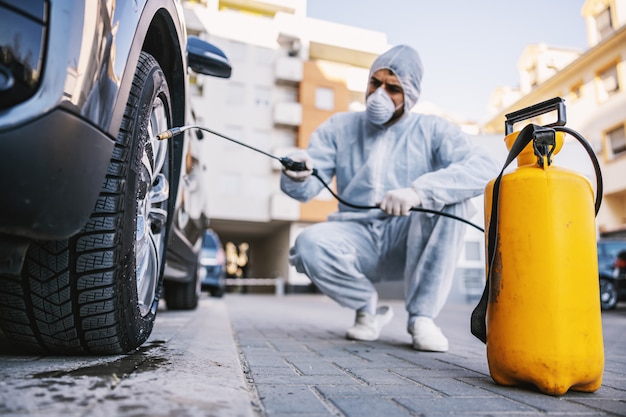 Homme en tenue de protection avec masque désinfectant les pneus de voiture, prévient l'infection par le virus Covid-19 covid-19, la contamination de germes ou de bactéries. Prévention des infections et contrôle de l'épidémie. Protection su