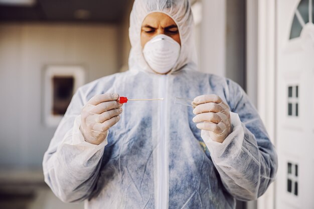 Homme en tenue de protection avec masque tenant un échantillon de coton-tige buccal et tube à essai prêt à collecter l'ADN des cellules. Coronavirus (COVID-19. épidémie. Pandémie mondiale