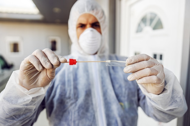 Homme en tenue de protection avec masque tenant un échantillon de coton-tige buccal et tube à essai prêt à collecter l'ADN des cellules. Coronavirus (COVID-19. épidémie. Pandémie mondiale