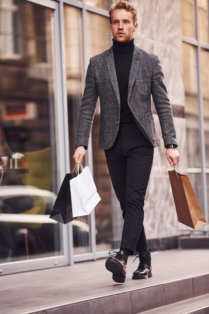 L'homme en tenue de soirée élégante et avec des forfaits shopping est à l'extérieur contre un bâtiment moderne.