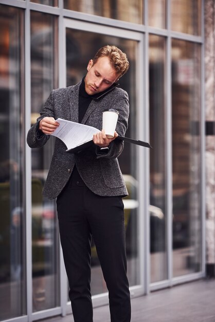 Un homme en tenue de soirée élégante avec une tasse de boisson et un bloc-notes dans les mains est à l'extérieur contre un bâtiment moderne et a une conversation par téléphone.