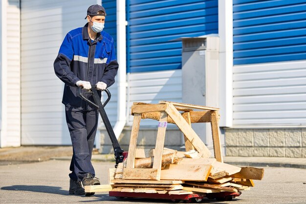 Un homme en tenue de travail et un masque médical porte une charge sur un chariot sur le territoire d'une base industrielle un jour d'été