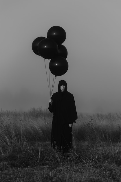 Un homme terrible dans une cape à capuchon avec des ballons à la main se dresse dans un champ brumeux. Concept sombre de cauchemars mystiques