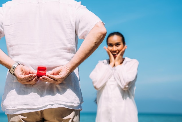 Un homme tient une bague de mariage à sa petite amie étonnée sur la plage avec le soleil