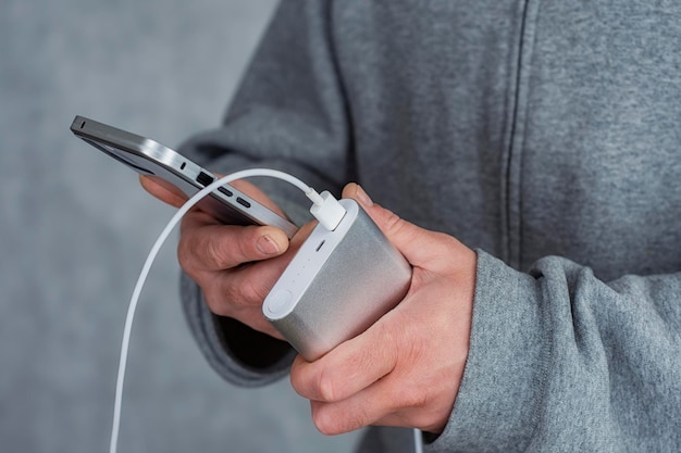 Un homme tient une banque d'alimentation dans ses mains et charge un smartphone sur fond gris.