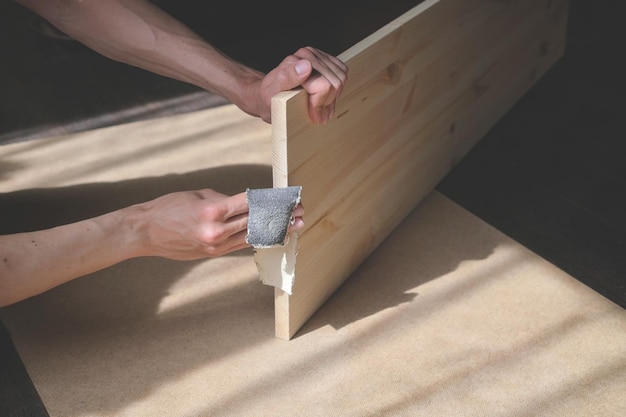L'homme tient une planche de bois avec ses mains pour polir le bord avec du papier de verre pour fabriquer des meubles faits à la main
