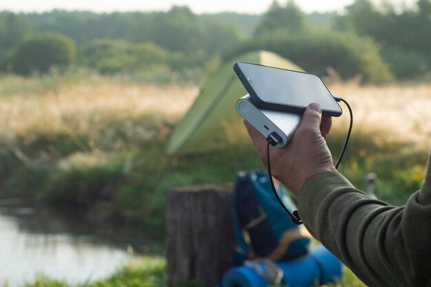 L'homme tient un smartphone dans ses mains et le charge avec une banque d'alimentation dans le contexte d'une tente touristique dans la nature. Chargeur de voyage portable.