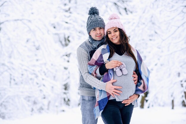 L'homme tient le ventre de femme enceinte et la femme tient les chaussures de bébé dans sa main alors qu'ils se tiennent sur le parc d'hiver enneigé