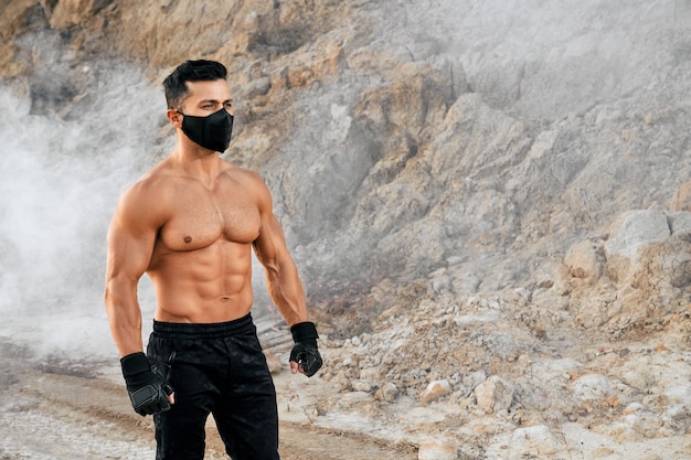 Homme torse nu athlétique portant un masque de protection et un pantalon de sport montrant son corps musclé en se tenant debout dans une carrière de sable