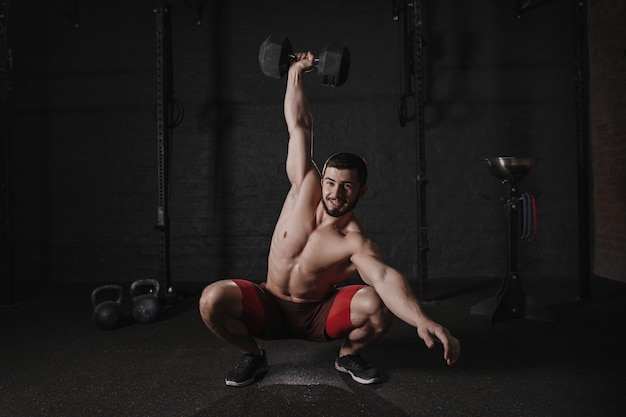 Homme torse nu faisant des squats d'haltères et souriant. Bel athlète crossfit faisant un entraînement fonctionnel.