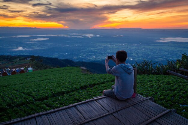 Homme de tourisme assis sur une colline pittoresque et prend une photo de la vallée du matin par smartphone.