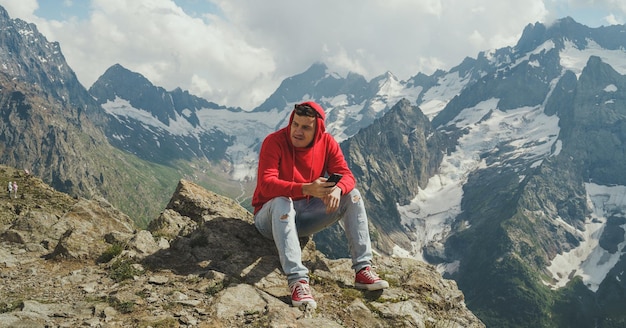 Homme touriste assis sur un rocher avec un smartphone dans les montagnes Voyageur masculin utilisant un téléphone portable contre un ciel nuageux par une journée ensoleillée en terrain montagneux