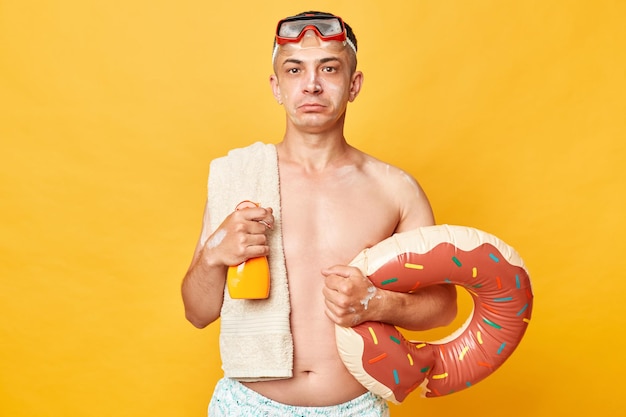 Homme touriste sérieux dans une station balnéaire debout avec un anneau en caoutchouc isolé sur fond jaune tenant un écran solaire regardant la caméra avec une expression confiante