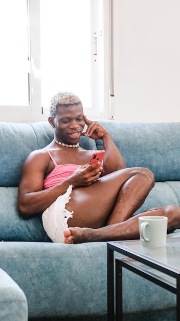 Homme transsexuel noir africain dans des vêtements féminins élégants envoyant des SMS sur un téléphone portable sur un canapé